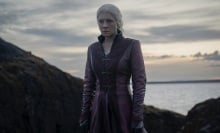 Rhaenyra from "House of the Dragon" stands on a rocky cliff by the sea, wearing a long red coat.