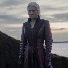 Rhaenyra from "House of the Dragon" stands on a rocky cliff by the sea, wearing a long red coat.