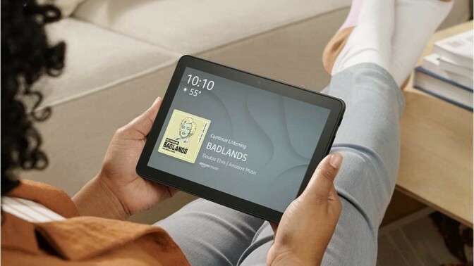 a person sits with legs extended on a coffee table while holding the fire HD 8 tablet in their hands. The display screen of the tablet shows the time, temperature, and the current song playing