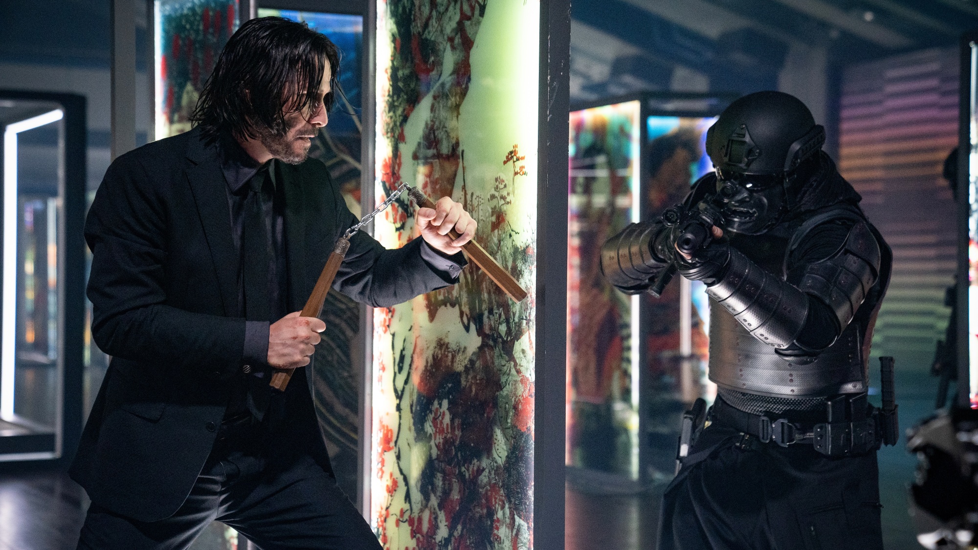 A man in a black suit wields nunchuks against a gun-toting man in black tactical armor.