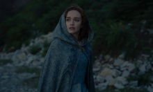 Alicent Hightower stands outside on the island of Dragonstone, wearing a blue dress and cloak.