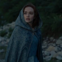 Alicent Hightower stands outside on the island of Dragonstone, wearing a blue dress and cloak.