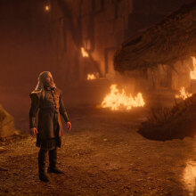 A bearded man stands in a gloomy cave, looking up at a dragon.