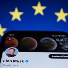 Elon Musk's bluecheck account on a phone against a European Union flag. 