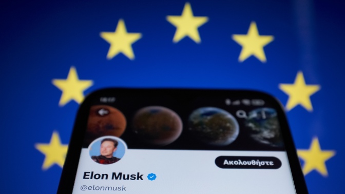 Elon Musk's bluecheck account on a phone against a European Union flag. 