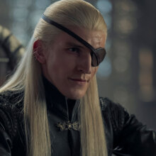 A man with long blonde hair and an eyepatch sits on a chair.