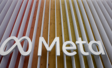 The Meta logo sign in front of a building. The log is white, the building is covered in white lines, interspersed with red, orange, and yellow lines.