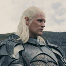 A man with long blonde hair in armour stands on a hillside.