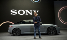 An asian man in a suit stands in front of a car. In the background is a giant screen with the words Sony displayed