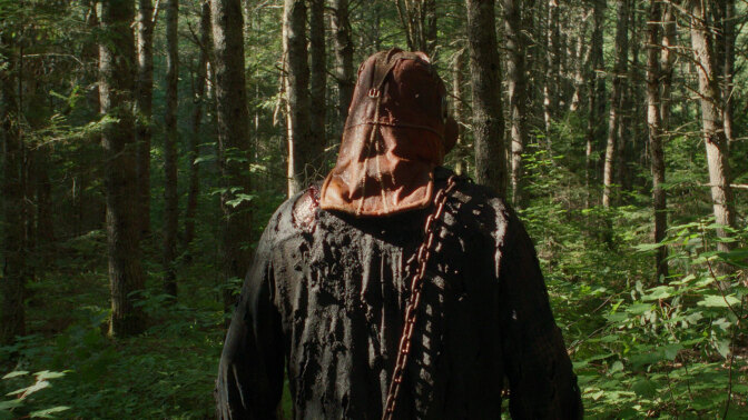 A man wearing a mask is seen from behind as he walks through the woods.