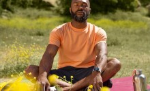 a person wearing an orange shirt and black shorts sits cross-legged in a field of grass while wearing the google pixel 2 watch on his left wrist.