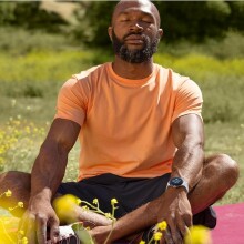 a person wearing an orange shirt and black shorts sits cross-legged in a field of grass while wearing the google pixel 2 watch on his left wrist.