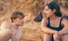 A young man and young woman sit on the beach looking romantically at each other.