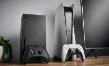 An Xbox Series X and PlayStation 5 sitting side by side next to a TV, with a console-appropriate controller propped up against each one.