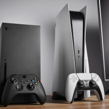 An Xbox Series X and PlayStation 5 sitting side by side next to a TV, with a console-appropriate controller propped up against each one.