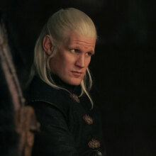 A blonde man in medieval clothes sits in a dark room.