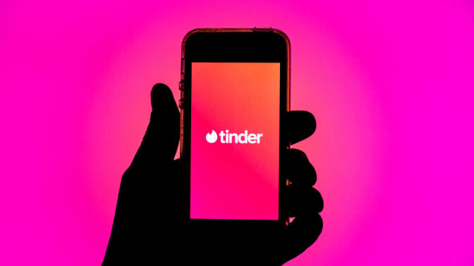 Silhouette of a hand holding a smartphone displaying the Tinder app.