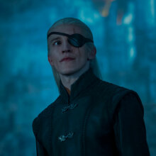 A man with blonde hair and an eyepatch stands in a gloomy room.