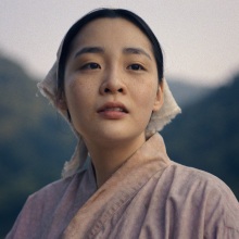 A close-up of young Sunja in the Japanese countryside, with large forested hills behind her.