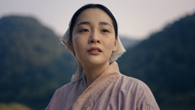 A close-up of young Sunja in the Japanese countryside, with large forested hills behind her.