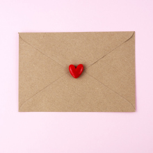 A brown envelope with a red heart on top of it, on a pale pink background. 