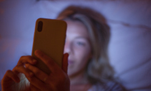 A white woman with blonde hair lies in bed at night scrolling on her phone. 
