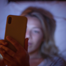 A white woman with blonde hair lies in bed at night scrolling on her phone. 