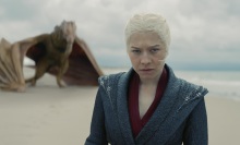 Rhaenyra Targaryen stands on a beach with her yellow dragon Syrax behind her.