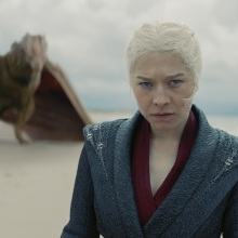 Rhaenyra Targaryen stands on a beach with her yellow dragon Syrax behind her.