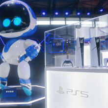 Two PS5 consoles in a glass display case next to a large Astro Bot statue