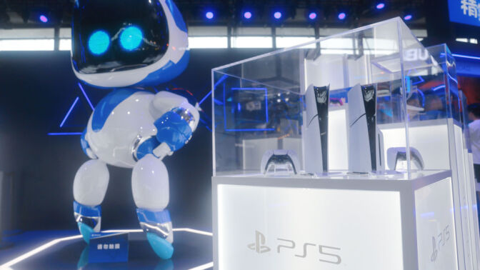 Two PS5 consoles in a glass display case next to a large Astro Bot statue