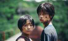 Two kids covered in mud stand on a railway track in a forest, looking at the camera.