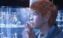 A man with orange hair admires a golden oyster earring in a jewelry display case.