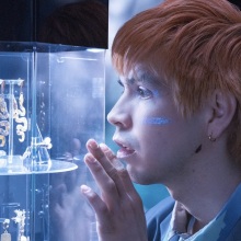 A man with orange hair admires a golden oyster earring in a jewelry display case.