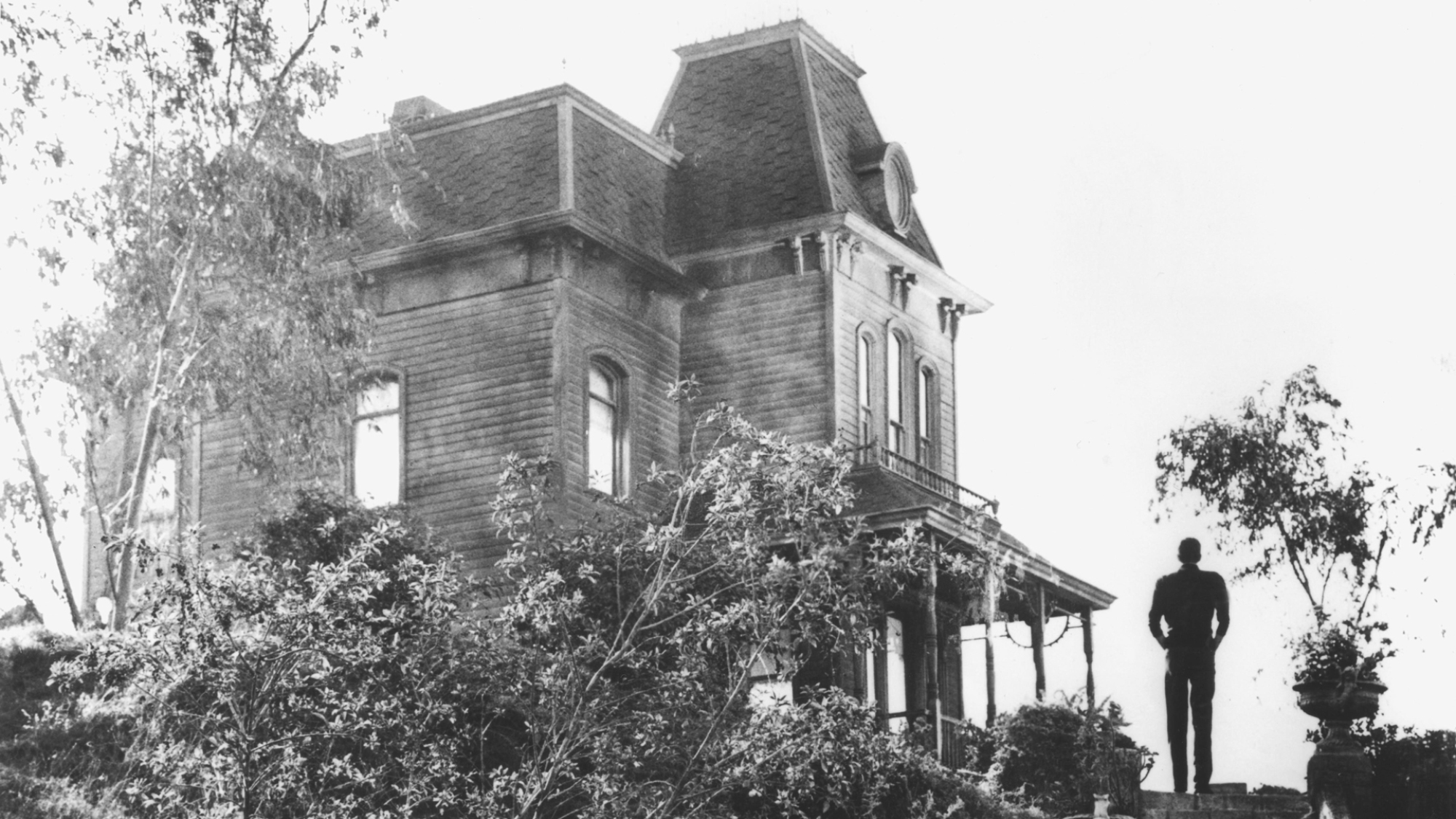 A black and white film still from Alfred Hitchcock's "Psycho" showing an old house on a hill with the silhouette of a man.
