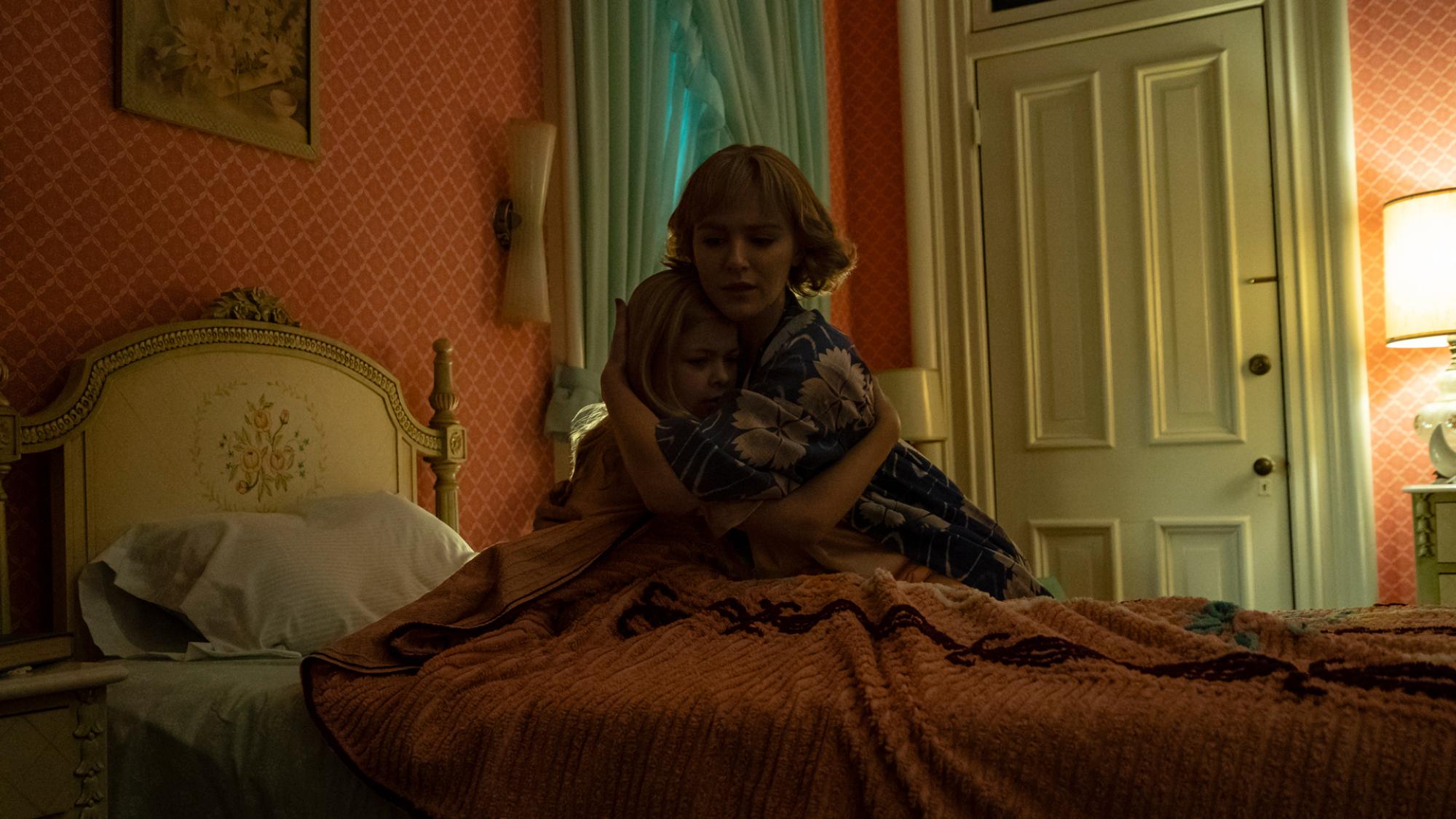 A woman and her daughter hug in a bedroom with pink wallpaper and antique furniture.