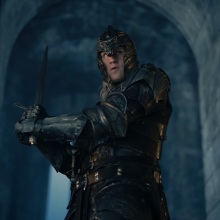 Daemon Targaryen walks through a large doorway in Harrenhal, holding a sword and wearing full armor.