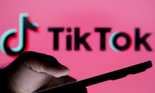 A hand holding a phone in front of a blurry sign reading TikTok on a pink backdrop.