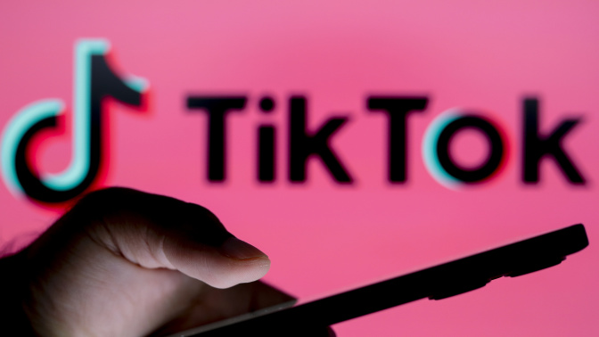 A hand holding a phone in front of a blurry sign reading TikTok on a pink backdrop.