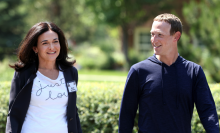 Sheryl Sandberg in a white graphic tee and navy blazer smiles and walks beside Zuckerberg in a navy pullover.