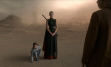 A woman with a sword on her back stands next to a little girl in the desert.