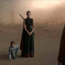 A woman with a sword on her back stands next to a little girl in the desert.