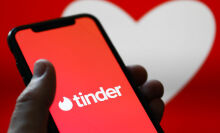 Tinder logo displayed on a phone screen and a heart shape displayed on a screen in the background.