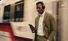 a male wearing the sonos ace stands on a train platform as the train whizzes by behind him