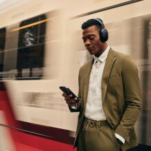 a male wearing the sonos ace stands on a train platform as the train whizzes by behind him