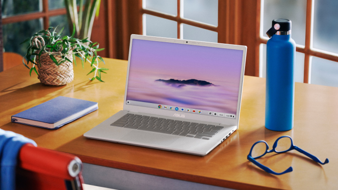 the asus chromebook plus cx34 on a wooden desk in front of a window next to a notebook, a water bottle, and a pair of glasses