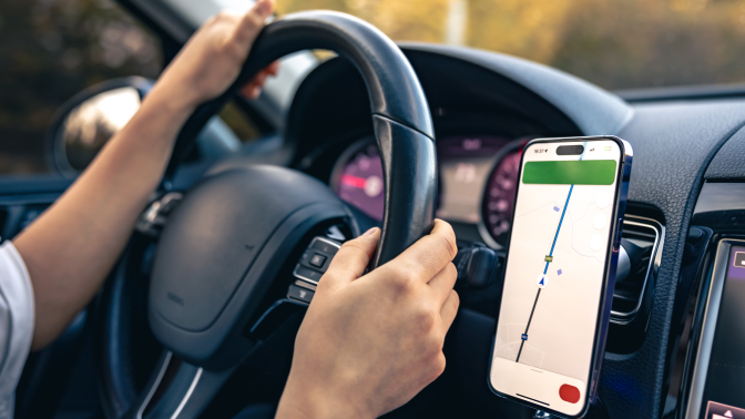 A person's hands on a car steering wheel. A phone with GPS navigation on the screen is attached to the dashboard.