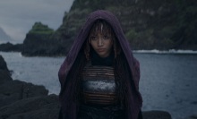 Mae from "The Acolyte" stands on a rock by the ocean, a purple cloak covering her head.