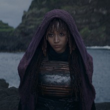 Mae from "The Acolyte" stands on a rock by the ocean, a purple cloak covering her head.