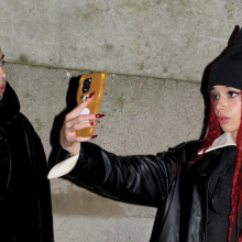 Two young women on a sidewalk, one talking on her cell phone, the other taking a selfie while wearing a black hat with cat ears